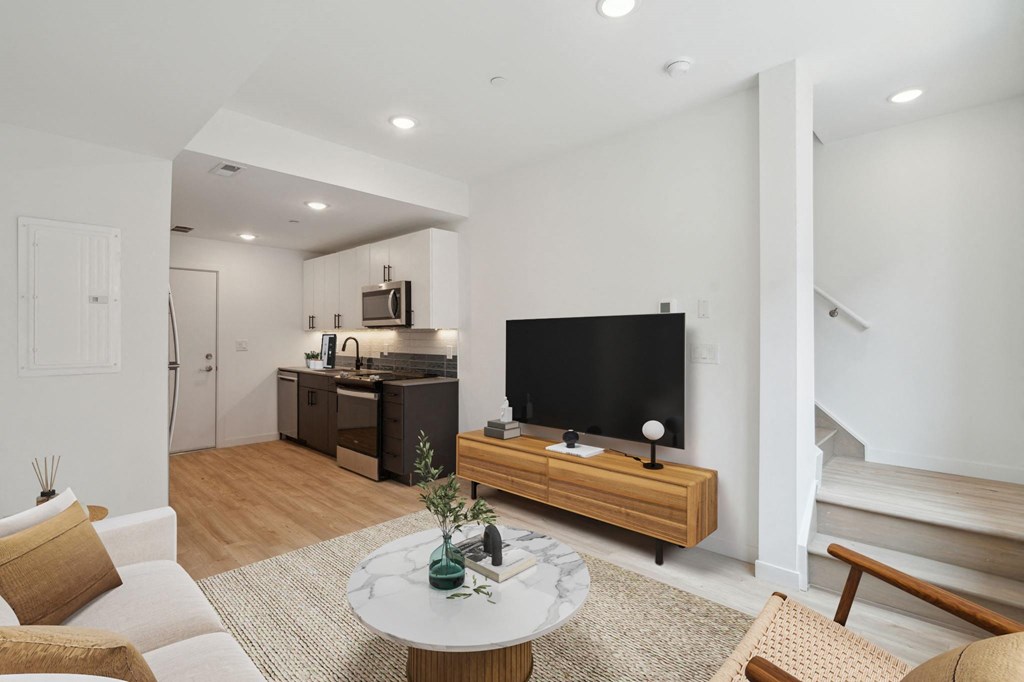 Modern open-plan living space with hardwood flooring, featuring a cozy sitting area with a white round marble-top coffee table, sleek TV stand, and an understairs white door.