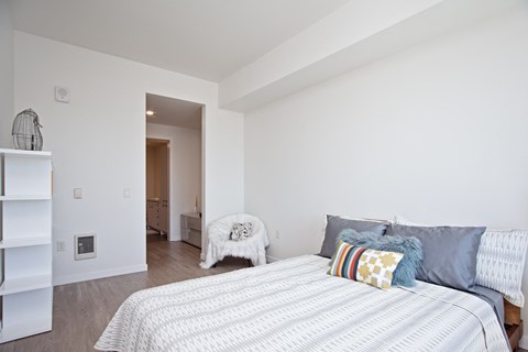 Beaverton, OR, Apartments with In-Unit Washer and Dryer - Westline - Bright Bedroom With White Bedding, Wood Style Flooring, Open Shelving, And Nearby Bathroom Entry In A Modern Apartment.