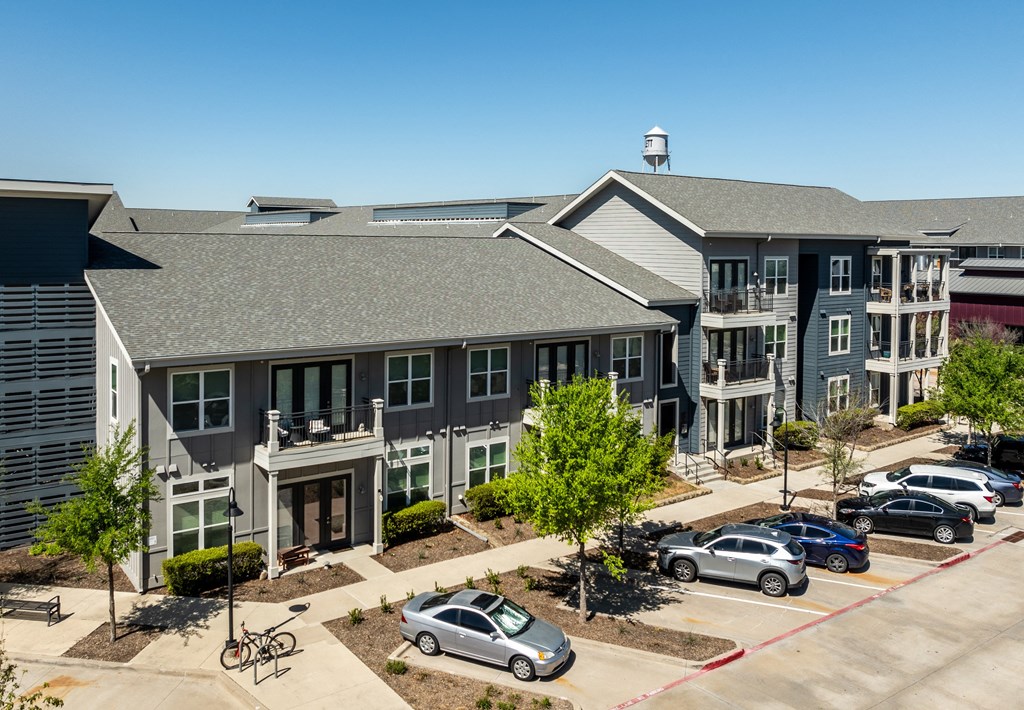 Apartments for Rent Rowlett, TX - Village of Rowlett - Aerial View of Property Exterior with Grey and Blue Walls, Showcasing Nearby Trees and Parking Spaces.