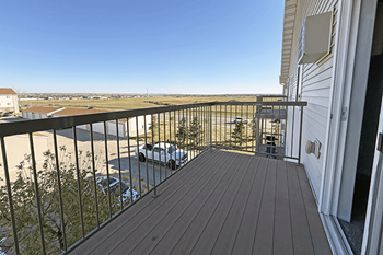 A balcony with a wooden floor and a metal railing overlooks a parking lot and distant buildings.
