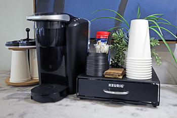A Keurig coffee maker is on a counter with a few cups and a plant.