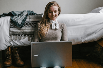 A woman in a grey sweater is using a laptop on a wooden floor.