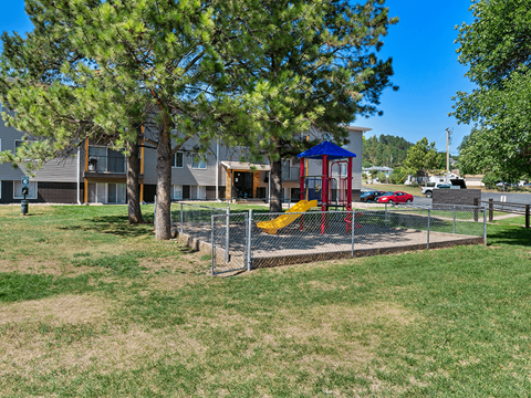 A playground with a red swing set and a blue roof is surrounded by a fence and a grassy area.