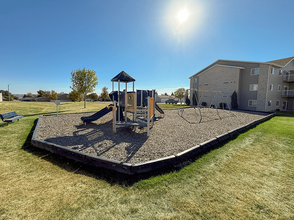A playground with a swing set and a slide in the foreground and apartment buildings in the background.