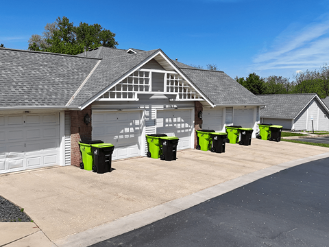 A white building with a black roof and a row of green bins in front.