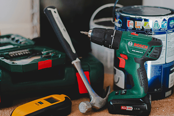 A green Bosch drill is on a carpeted floor next to a hammer and a yellow and black tape measure.