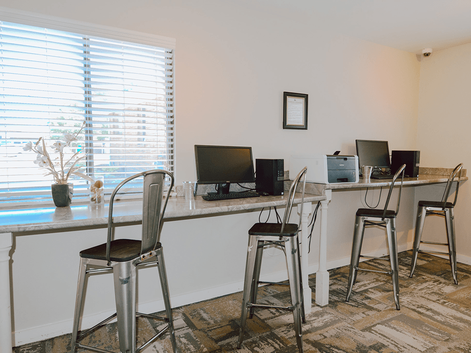 two computers on a long desk with three stools in a room with a large window