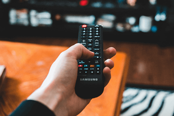 A hand is holding a Samsung remote control in front of a television.