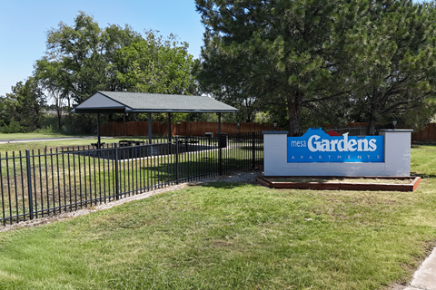 A sign for Mega Gardens stands in front of a fence and trees.