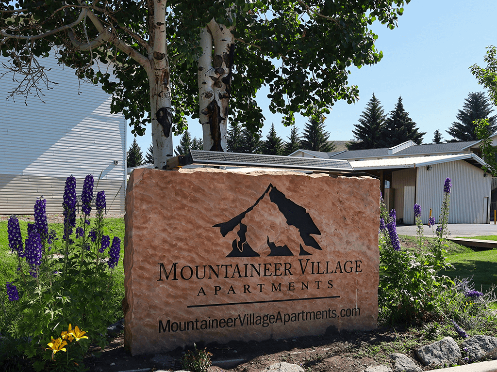 A sign for Mountaineer Village Apartments is surrounded by flowers and trees.