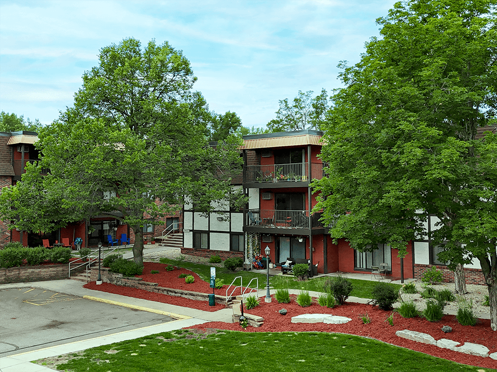 A red building with a white trim and a balcony is surrounded by trees and bushes.
