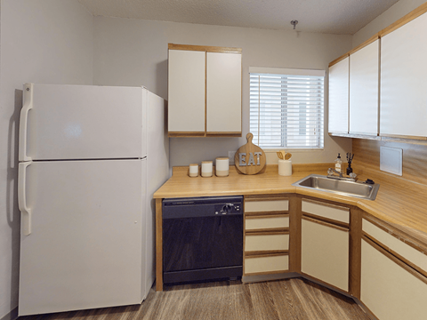 A white refrigerator stands in a kitchen with wooden cabinets and a black dishwasher.
