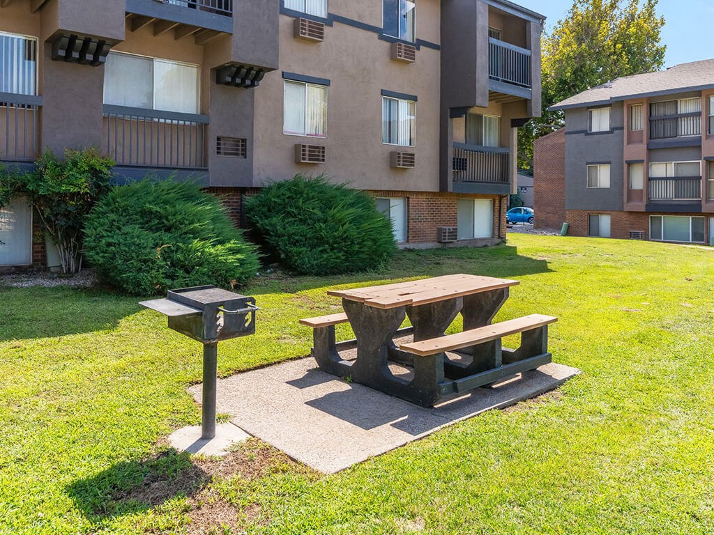 A wooden picnic table sits in the grass in front of apartment buildings.