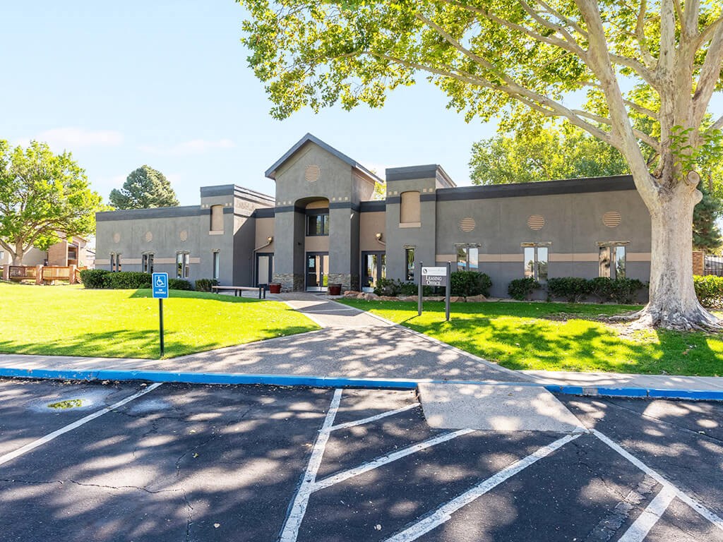 A parking lot in front of a grey building with a tree on the right.