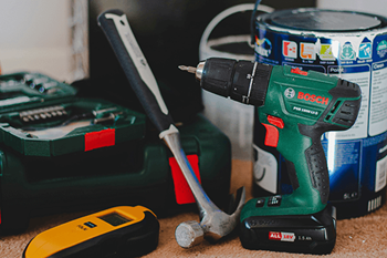A green Bosch drill is on a carpeted floor next to a hammer and a yellow and black device.