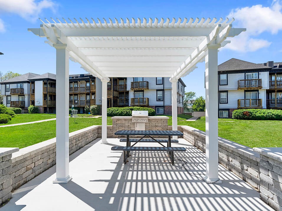 a picnic table under a white pergola in front of an apartment building