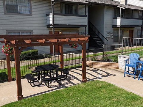 A wooden pergola with a black table and chairs is in front of a house.