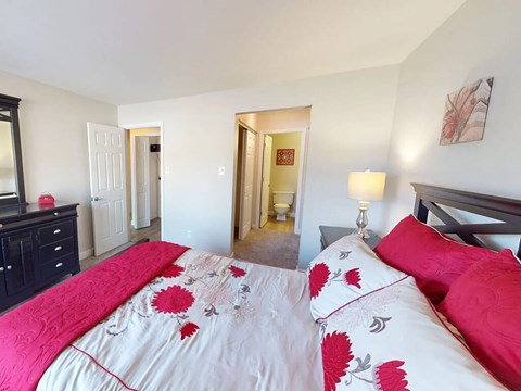 A bed with a red and white comforter is in the foreground of a bedroom.