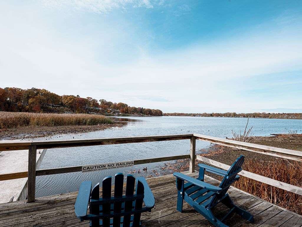 Two blue chairs are on a wooden deck overlooking a body of water.