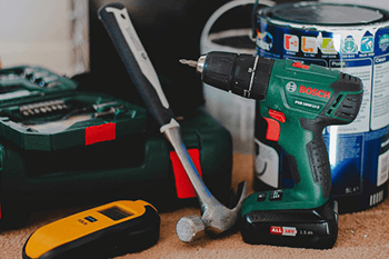 A green and black power drill is on a carpeted floor next to a yellow and black tape measure and a hammer.
