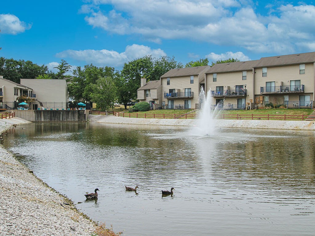 a fountain in a pond with ducks in front of an apartment building