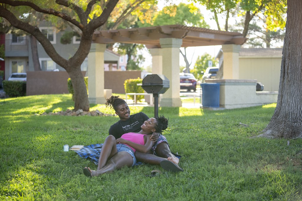 Two people are sitting on the grass under a tree. at Solstice Apartment Homes, Las Vegas, NV