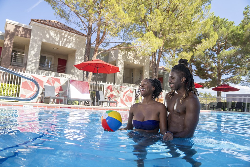 A couple is sitting in a pool with a ball in the foreground. at Solstice Apartment Homes, Las Vegas, 89108