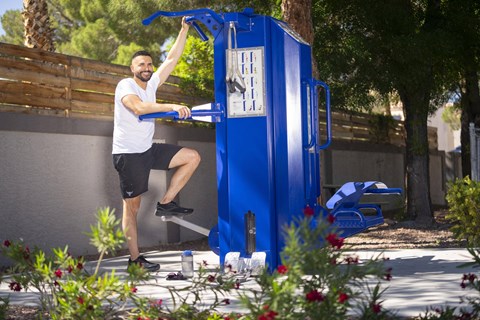 A man in a white shirt and black shorts is exercising on a blue gym machine outdoors. at The 95 Apartments, Las Vegas