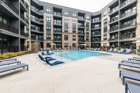 A large outdoor pool surrounded by lounge chairs and umbrellas in front of apartment buildings.