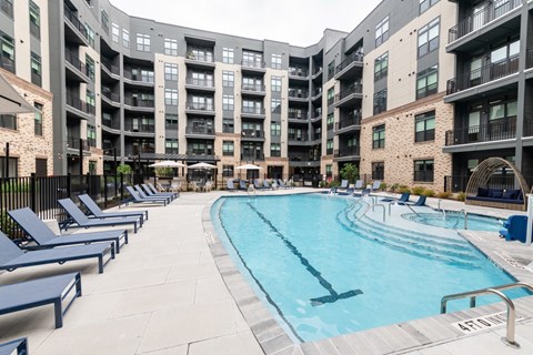 A large swimming pool surrounded by lounge chairs in front of apartment buildings.