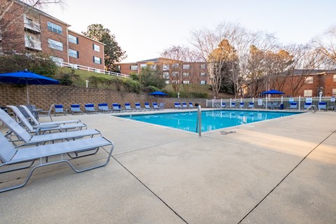 A pool surrounded by chairs and umbrellas with apartment buildings in the background at Amelia Village apartments in Clayton, NC.