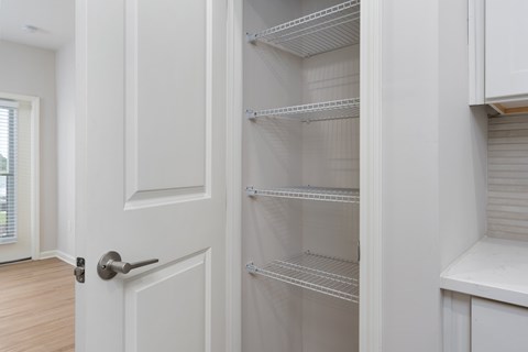 A white door with a handle is open to a white pantry with empty shelves at Evolve Holly Ridge Apartments in Holly Ridge, NC.