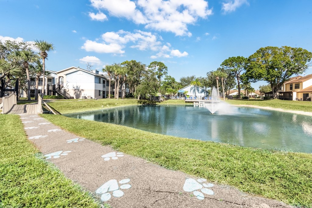 a pathway leading to a pond with a fountain and houses in the background