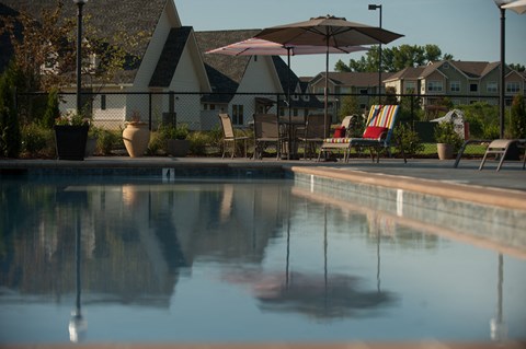 A pool with a reflection of a building and a patio with chairs and umbrellas.