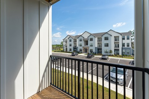 A view of a balcony with a black railing and a white wall on the left side, overlooking a parking lot with cars and a building in the distance. at Evolve Holly Ridge Apartments in Holly Ridge, NC