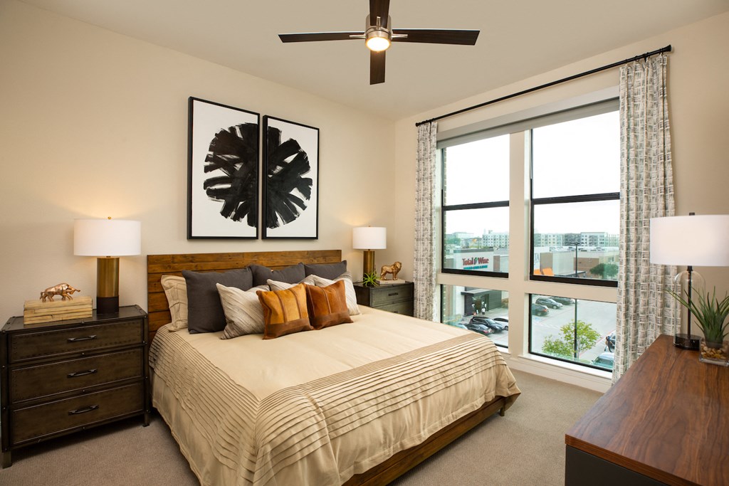 A bedroom with a large bed, a ceiling fan, and a view of the city at 27Seventy Lower Heights apartments in Houston, TX.