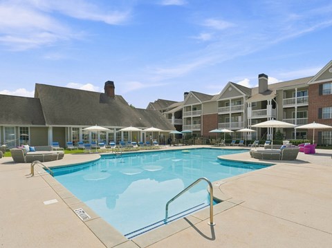 A large swimming pool surrounded by lounge chairs and umbrellas in front of apartment buildings.