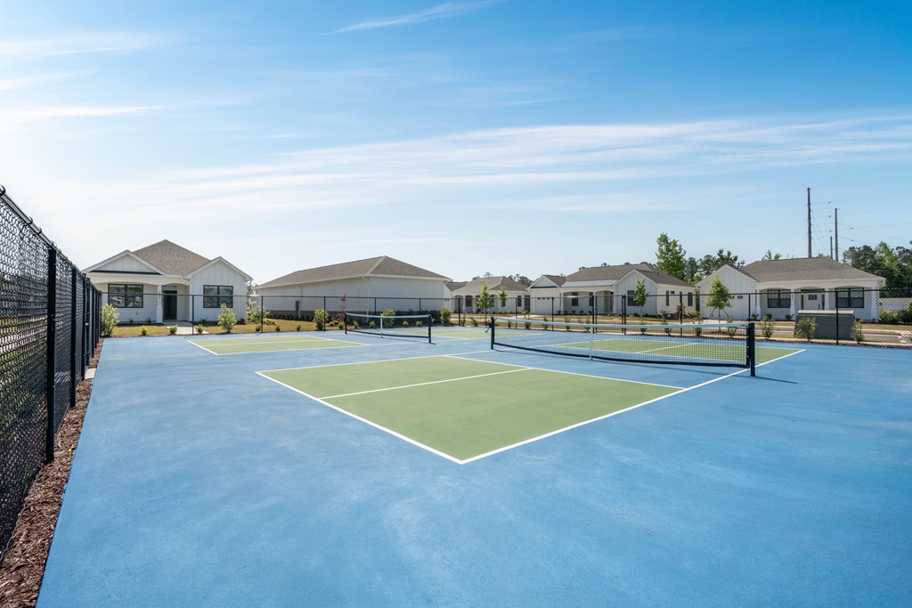 A tennis court is surrounded by a fence and there are houses in the background.