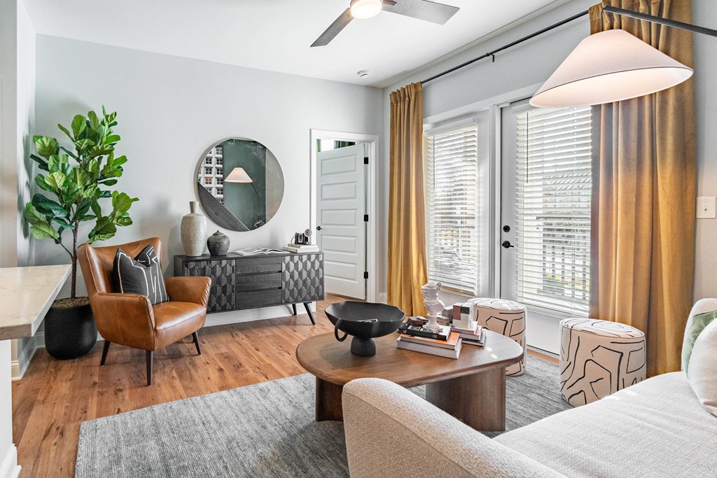 A living room with a grey rug, a white sofa, a wooden coffee table, a round mirror, a ceiling fan, and a potted plant.