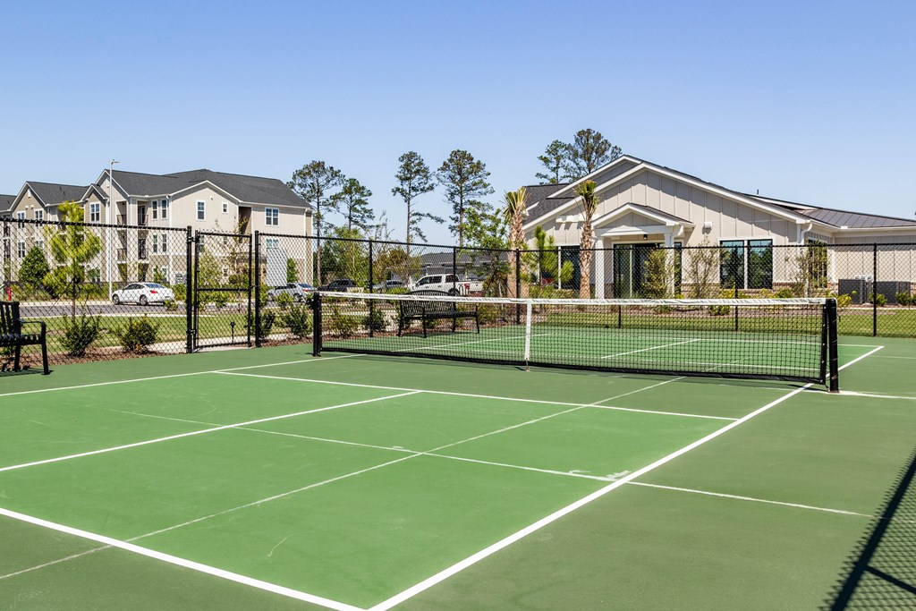 A tennis court is surrounded by a fence and a building. at Evolve Waterford Apartments in Belville, NC