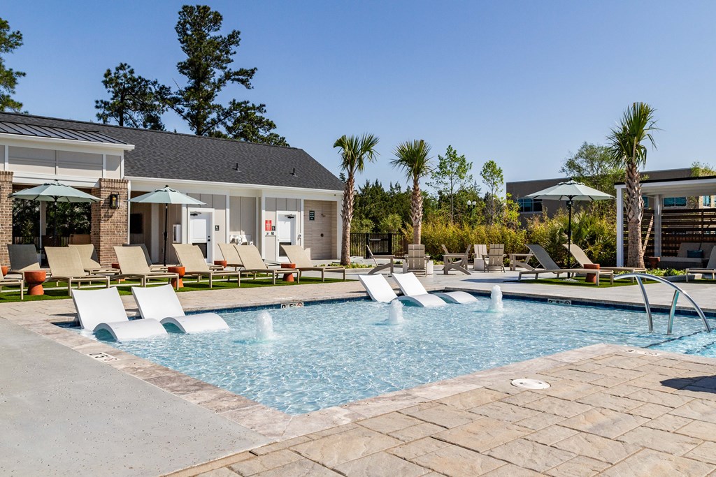A pool with a waterfall and a patio with chairs and umbrellas at Evolve Waterford Apartments in Belville, NC