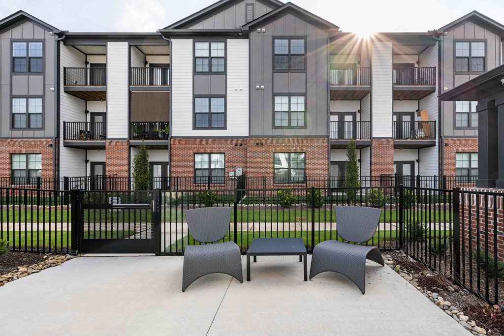 Two grey chairs are placed on a concrete walkway in front of a black fence and apartment building.