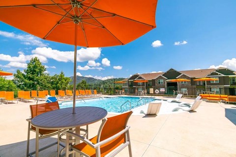 A large orange umbrella is shading a table and chairs by a pool.