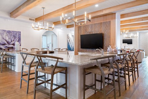 A bar with a counter and chairs is in the foreground of a room with a television and a painting on the wall.