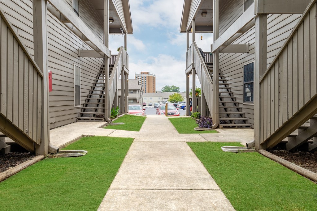 a path between two buildings with grass and a street in the distance