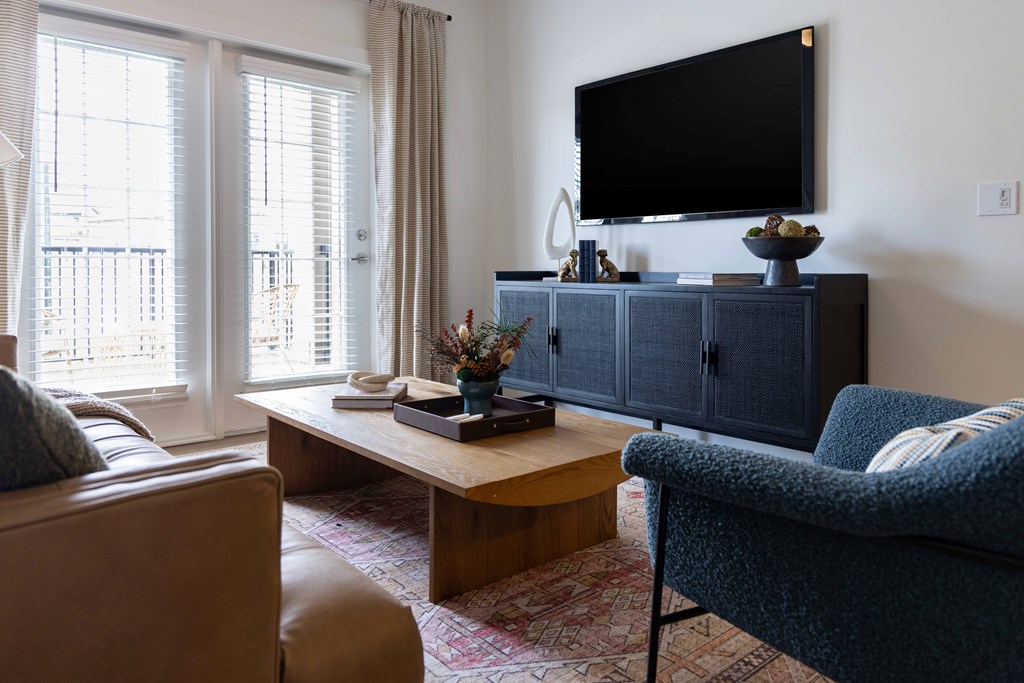 Interior living room with a brown leather couch, a blue armchair, a wooden coffee table, and a flat-screen TV mounted on the wall.