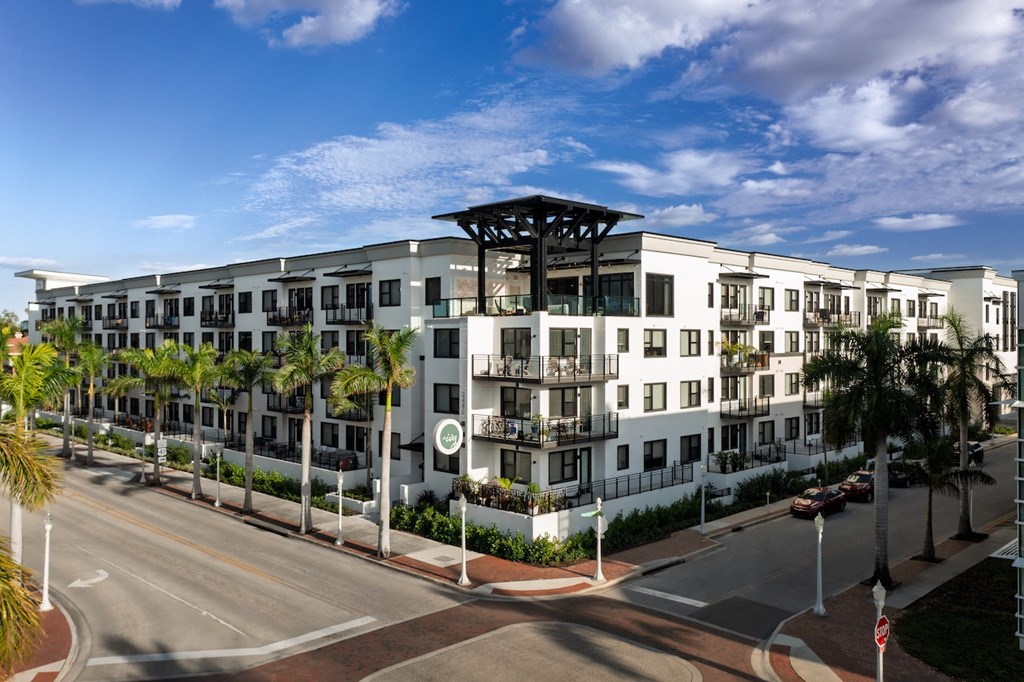 A modern white building with balconies and a black roof is surrounded by palm trees.