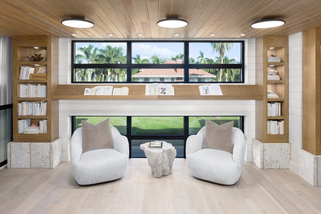 A room with two white chairs and a table in front of a window with a view of a lawn and trees.