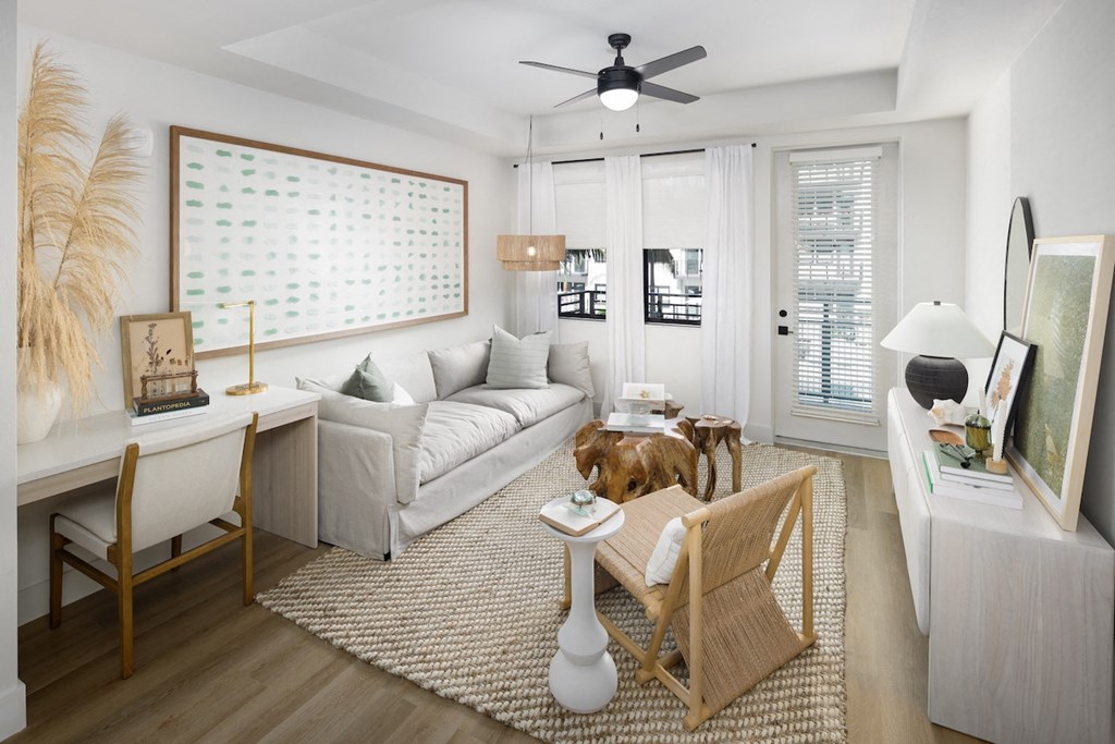 A living room with a white couch, a wooden chair, a white coffee table, a white rug, a white floor lamp, a white ceiling fan, a white wall with a calendar, a white door, a white window with blinds