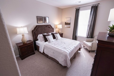 A bedroom with a large bed, a nightstand with a lamp, a chair, and a window with curtains.